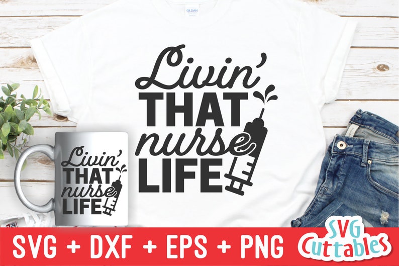 Pu&ograve; includere: Una maglietta bianca con testo nero che recita "Livin' THAT nurse LIFE" con un'immagine di una siringa. La maglietta &egrave; esposta su una superficie di legno con una tazza bianca e jeans blu.