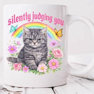 Pu&ograve; includere: Tazza in ceramica bianca con un gattino grigio con un'espressione imbronciata. La tazza presenta un design floreale con rose e margherite rosa, un arcobaleno e la scritta "silently judging you". Sono incluse anche farfalle.