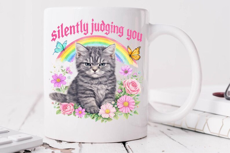 Pu&ograve; includere: Tazza in ceramica bianca con un gattino grigio con un'espressione imbronciata. La tazza presenta un design floreale con rose e margherite rosa, un arcobaleno e la scritta "silently judging you". Sono incluse anche farfalle.