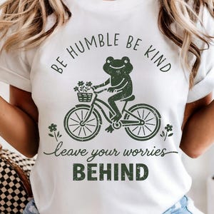 May include: White t-shirt featuring a green frog on a bicycle, carrying flowers. The text above reads "BE HUMBLE BE KIND", with "leave your worries BEHIND" below. A casual, nature-themed design.