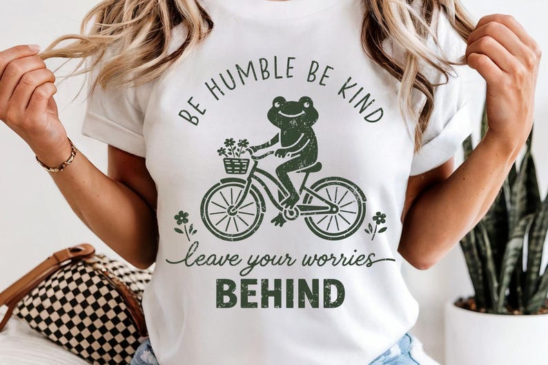 May include: White t-shirt featuring a green frog on a bicycle, carrying flowers. The text above reads "BE HUMBLE BE KIND", with "leave your worries BEHIND" below. A casual, nature-themed design.