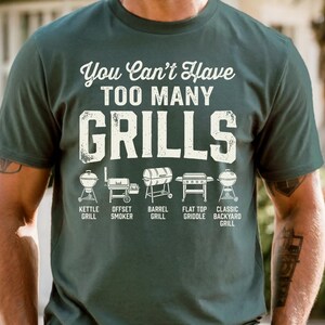May include: Dark green t-shirt with the text "You Can't Have TOO MANY GRILLS" in white. Below the text are illustrations of different grill types: kettle, offset smoker, barrel, flat top griddle, and classic backyard grill.