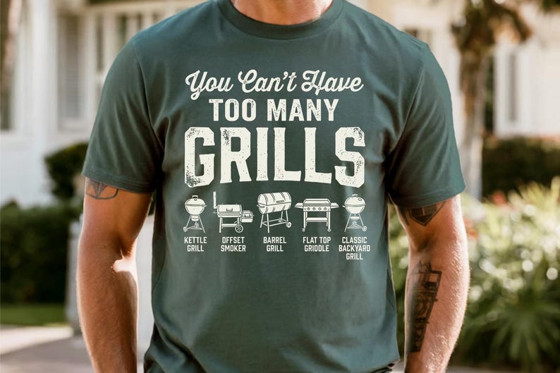 May include: Dark green t-shirt with the text "You Can't Have TOO MANY GRILLS" in white. Below the text are illustrations of different grill types: kettle, offset smoker, barrel, flat top griddle, and classic backyard grill.