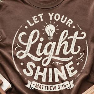 May include: Brown t-shirt with the phrase "Let Your Light Shine" in a circular design. The design includes a lightbulb illustration and the Bible verse Matthew 5:16. The shirt is a casual wear item.