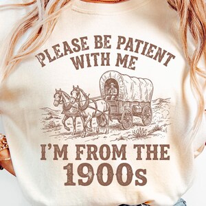 May include: Cream-colored t-shirt featuring a sepia-toned illustration of a covered wagon pulled by two horses. The text above reads "PLEASE BE PATIENT WITH ME" and below, "I'M FROM THE 1900s".