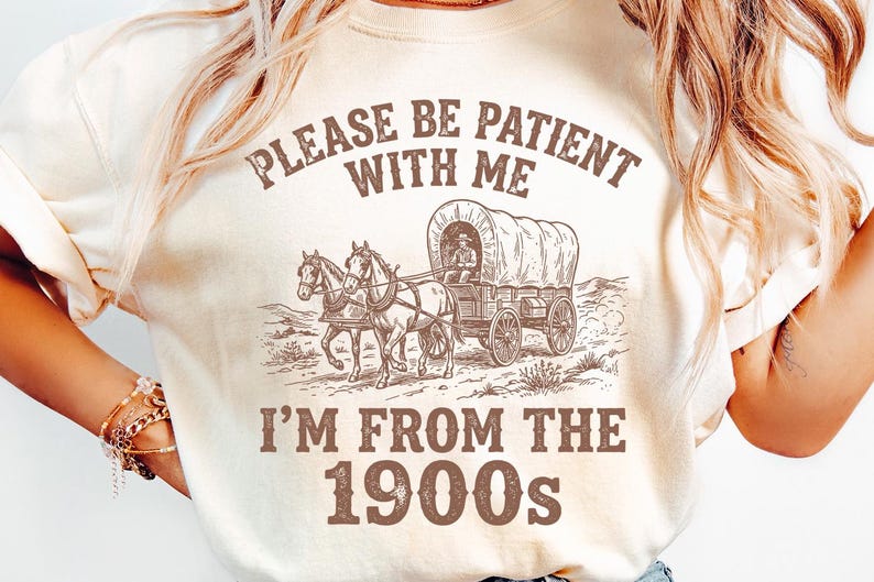 May include: Cream-colored t-shirt featuring a sepia-toned illustration of a covered wagon pulled by two horses. The text above reads "PLEASE BE PATIENT WITH ME" and below, "I'M FROM THE 1900s".