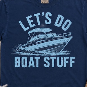 May include: A navy blue short-sleeve t-shirt featuring a light blue graphic of a motorboat on the water. The text "LET'S DO BOAT STUFF" is printed in a distressed light blue font above and below the boat illustration on the front of the shirt.