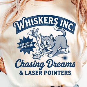 Puede incluir: Camiseta color crema con un gráfico de estilo vintage. El diseño incluye un gato juguetón con un juguete de plumas, un ratón y el texto "Whiskers Inc. Chasing Dreams & Laser Pointers". También hay una insignia de "Curiosity Approved".