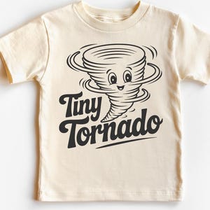 Peut inclure: T-shirt cr&egrave;me avec un dessin de tornade souriante en noir. Le texte "Tiny Tornado" est &eacute;crit en dessous. Le t-shirt a des manches courtes et un col rond. Id&eacute;al pour les enfants.