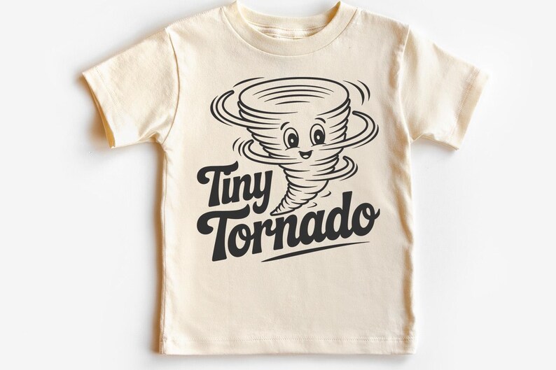 Peut inclure: T-shirt cr&egrave;me avec un dessin de tornade souriante en noir. Le texte "Tiny Tornado" est &eacute;crit en dessous. Le t-shirt a des manches courtes et un col rond. Id&eacute;al pour les enfants.