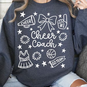 May include: Dark gray sweatshirt with a white graphic design. The design includes the words "Cheer Coach", a megaphone, a bow, a pom-pom, a cheerleader uniform, a basketball, and the words "Go Team".