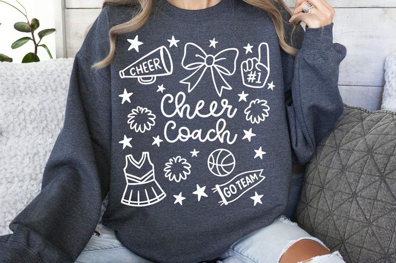 May include: Dark gray sweatshirt with a white graphic design. The design includes the words "Cheer Coach", a megaphone, a bow, a pom-pom, a cheerleader uniform, a basketball, and the words "Go Team".