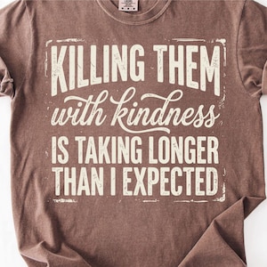 K&ouml;nnte beinhalten: Braunes T-Shirt mit dem Text "KILLING THEM with kindness IS TAKING LONGER THAN I EXPECTED" in verblichener wei&szlig;er Schrift. Das Shirt hat hochgekrempelte &Auml;rmel und einen Rundhalsausschnitt.