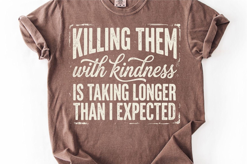 K&ouml;nnte beinhalten: Braunes T-Shirt mit dem Text "KILLING THEM with kindness IS TAKING LONGER THAN I EXPECTED" in verblichener wei&szlig;er Schrift. Das Shirt hat hochgekrempelte &Auml;rmel und einen Rundhalsausschnitt.