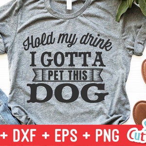 May include: A gray t-shirt with the text "Hold my drink I GOTTA PET THIS DOG" printed in black. The text is in a bold, blocky font. The shirt is a popular design for dog lovers.