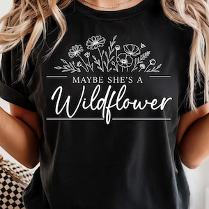 May include: Black t-shirt with white floral design and the text "MAYBE SHE'S A Wildflower". The shirt features a crew neck and short sleeves. The design includes various flowers and leaves.
