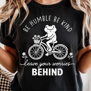 May include: Black t-shirt featuring a white graphic of a frog on a bicycle with a flower basket. The text says "BE HUMBLE BE KIND" and "leave your worries BEHIND".