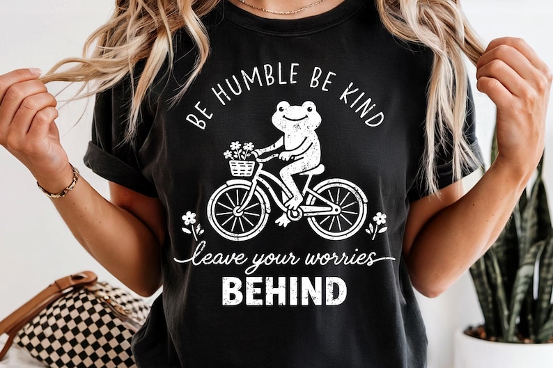 May include: Black t-shirt featuring a white graphic of a frog on a bicycle with a flower basket. The text says "BE HUMBLE BE KIND" and "leave your worries BEHIND".