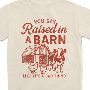 May include: Cream-colored t-shirt with a red graphic featuring a barn, cow, and chickens. The text reads "YOU SAY Raised in A BARN LIKE IT'S A BAD THING". A farm-themed design.