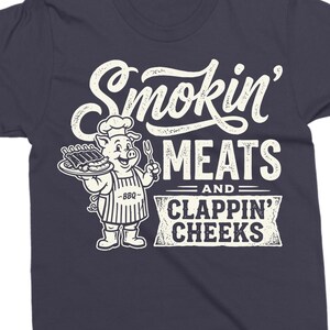 May include: Dark navy blue t-shirt with a white graphic. The graphic features the text "Smokin' Meats and Clappin' Cheeks" in a vintage style. A cartoon pig wearing a chef's hat and apron is holding a plate of ribs.