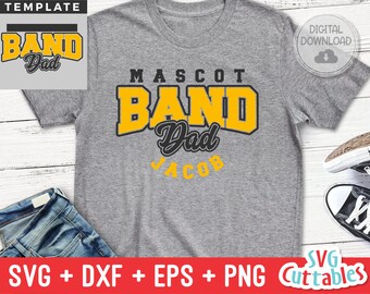 May include: A gray t-shirt with the text "MASCOT BAND Dad" in yellow and black lettering. The text "Jacob" is below the main text in black lettering. The t-shirt is on a white wooden surface.