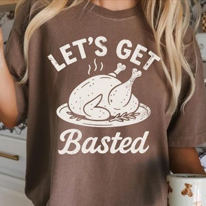 Puede incluir: Camiseta marrón con la frase "Let's Get Basted" y un gráfico de un pavo asado en un plato. El texto y el gráfico son blancos. La camiseta es de manga corta.