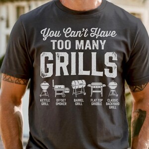May include: Dark gray t-shirt with the text "You Can't Have TOO MANY GRILLS" in white. Below the text are illustrations of different grill types: kettle, offset smoker, barrel, flat top griddle, and classic backyard grill.