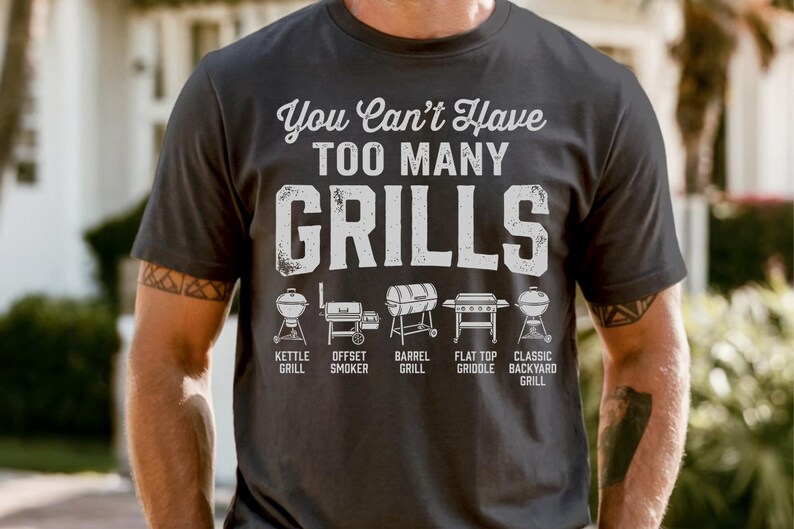 May include: Dark gray t-shirt with the text "You Can't Have TOO MANY GRILLS" in white. Below the text are illustrations of different grill types: kettle, offset smoker, barrel, flat top griddle, and classic backyard grill.