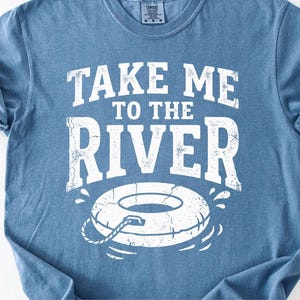 May include: Blue t-shirt with the text "TAKE ME TO THE RIVER" in distressed white lettering. Below the text is a graphic of a white inner tube with a rope. The shirt is made of a soft material.