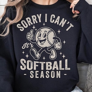 Op de afbeelding: Een marineblauwe sweatshirt met de witte tekst "SORRY I CAN'T It's SOFTBALL SEASON". De afbeelding toont een honkbalpersonage dat een duim omhoog geeft en loopt. Kleine witte sterren zijn verspreid rond het ontwerp.