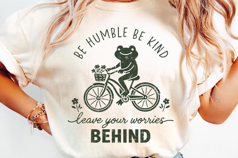 May include: A cream-coloured t-shirt featuring a dark green illustration of a frog on a bicycle, with a basket of flowers. The text reads "BE HUMBLE BE KIND" and "leave your worries BEHIND". A charming and positive design.