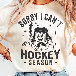 May include: A cream-coloured t-shirt featuring a black graphic of a hockey puck character with a hockey stick. The text says "SORRY I CAN'T It's HOCKEY SEASON". The design is in a vintage, cartoon style.