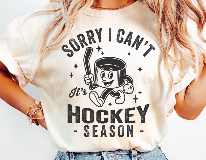 May include: A cream-coloured t-shirt featuring a black graphic of a hockey puck character with a hockey stick. The text says "SORRY I CAN'T It's HOCKEY SEASON". The design is in a vintage, cartoon style.