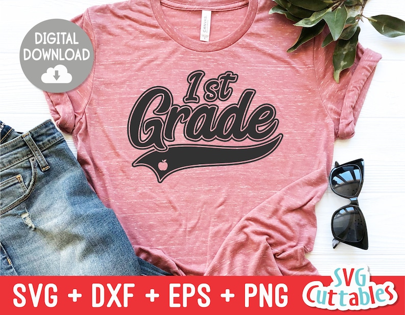 May include: A dusty rose t-shirt with the words "1st Grade" in a black, retro-style font. The design includes a small apple graphic. The shirt is paired with blue jeans and sunglasses, suggesting a back-to-school theme.