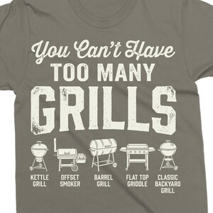 May include: A gray t-shirt with the text "You Can't Have Too Many Grills" in white. Below the text are illustrations of different grill types, including kettle, offset smoker, barrel, flat top griddle, and classic backyard grills.