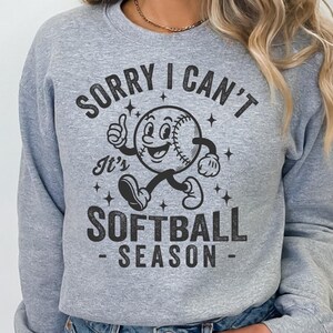 Op de afbeelding: Een lichtgrijs sweatshirt met de zwarte tekst "SORRY I CAN'T It's SOFTBALL SEASON". De afbeelding toont een softbalpersonage met een lachend gezicht, duim omhoog en lopende benen.