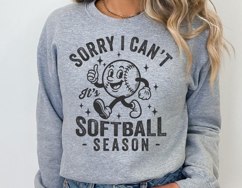 Op de afbeelding: Een lichtgrijs sweatshirt met de zwarte tekst "SORRY I CAN'T It's SOFTBALL SEASON". De afbeelding toont een softbalpersonage met een lachend gezicht, duim omhoog en lopende benen.