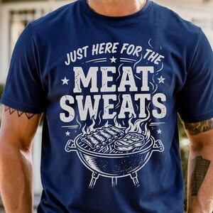 May include: Navy blue t-shirt with the text "JUST HERE FOR THE MEAT SWEATS" in white. A graphic of a grill with ribs and steak is below the text. The shirt is a casual wear item.