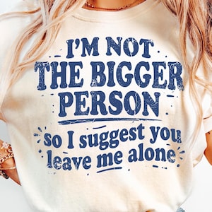 May include: A cream-coloured t-shirt featuring the text "I'M NOT THE BIGGER PERSON so I suggest you leave me alone" in distressed navy blue. The shirt has a crew neck and short sleeves.