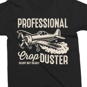 May include: Black t-shirt with a white graphic of a vintage crop duster airplane. The text "PROFESSIONAL Crop DUSTER" is above the plane, with "SILENT BUT DEADLY" below. The design has a distressed, vintage look.