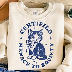 May include: Cream-colored sweatshirt with a navy blue graphic of a cat wearing a bow tie, surrounded by the text "CERTIFIED MENACE TO SOCIETY". The design includes stars. The sweatshirt is folded on a wooden surface.