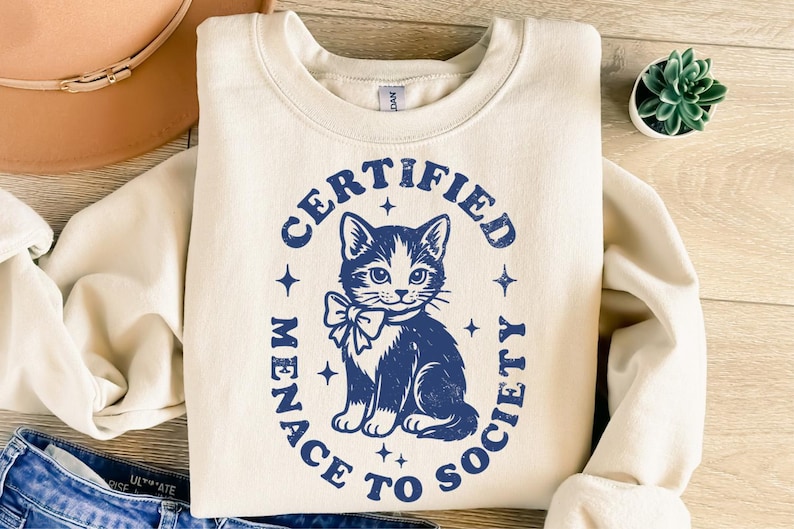 May include: Cream-colored sweatshirt with a navy blue graphic of a cat wearing a bow tie, surrounded by the text "CERTIFIED MENACE TO SOCIETY". The design includes stars. The sweatshirt is folded on a wooden surface.