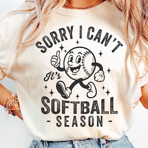 Op de afbeelding: Cr&egrave;mekleurig T-shirt met de zwarte tekst "SORRY I CAN'T It's SOFTBALL SEASON". De afbeelding toont een cartoon softbal met armen en benen, die een duim omhoog geeft. Het ontwerp bevat sterren.