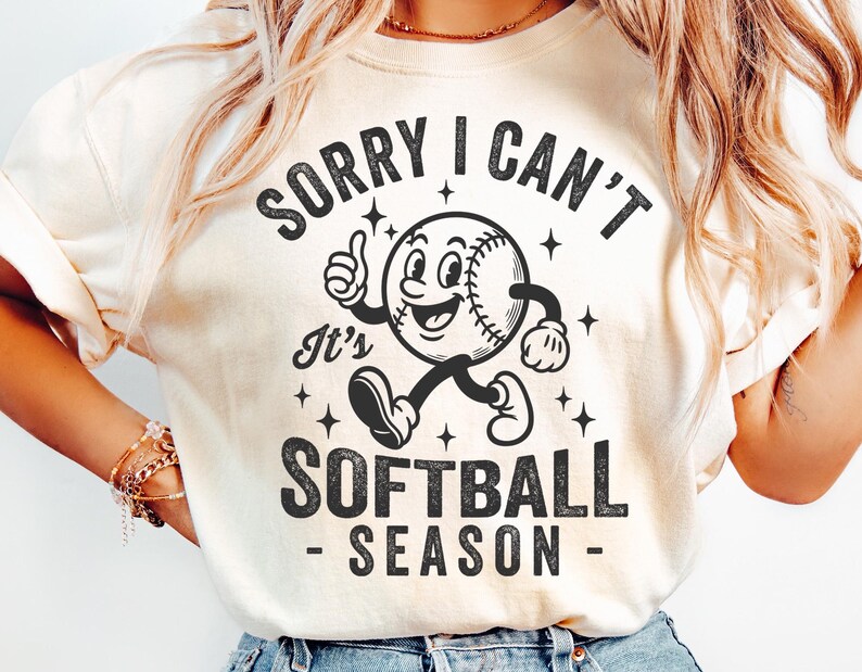 Op de afbeelding: Cr&egrave;mekleurig T-shirt met de zwarte tekst "SORRY I CAN'T It's SOFTBALL SEASON". De afbeelding toont een cartoon softbal met armen en benen, die een duim omhoog geeft. Het ontwerp bevat sterren.