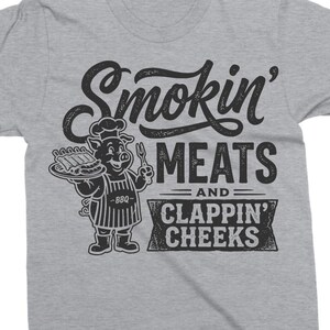 May include: A gray t-shirt with a black graphic. The graphic features the text "Smokin' MEATS and CLAPPIN' CHEEKS" with a cartoon pig wearing a chef's hat and apron, holding a plate of ribs and a fork.