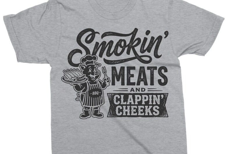 May include: A gray t-shirt with a black graphic. The graphic features the text "Smokin' MEATS and CLAPPIN' CHEEKS" with a cartoon pig wearing a chef's hat and apron, holding a plate of ribs and a fork.