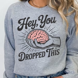 May include: A light gray sweatshirt with the text "Hey, You Dropped This" in black script. The graphic features a hand holding a pink brain, surrounded by radiating lines.
