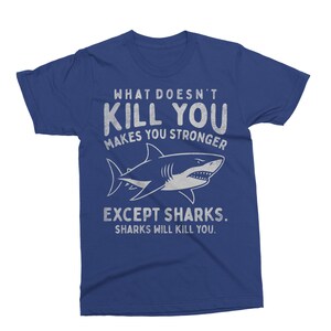 May include: A blue t-shirt with the text "WHAT DOESN'T KILL YOU MAKES YOU STRONGER EXCEPT SHARKS. SHARKS WILL KILL YOU." A white shark illustration is featured below the text. The shirt is a crew neck style.