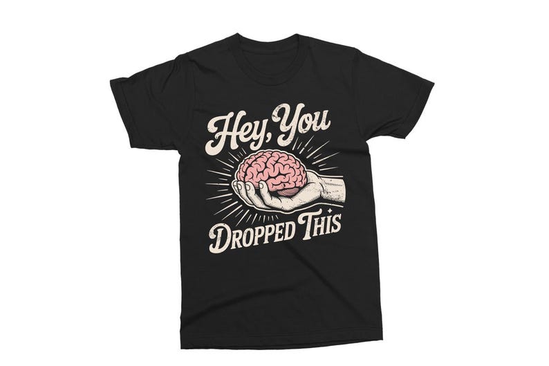 May include: Black t-shirt featuring a graphic design with the text "Hey, You Dropped This." The design includes a hand holding a pink brain, with radiating lines. The text and design are in white and pink.