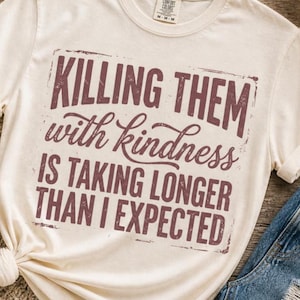 K&ouml;nnte beinhalten: Cremefarbenes T-Shirt mit dem Spruch "KILLING THEM with kindness IS TAKING LONGER THAN I EXPECTED" in einer verblassten, kastanienbraunen Schrift. Das Shirt ist aus weichem Material und hat kurze &Auml;rmel. Das Shirt wird mit blauen Jeans kombiniert.